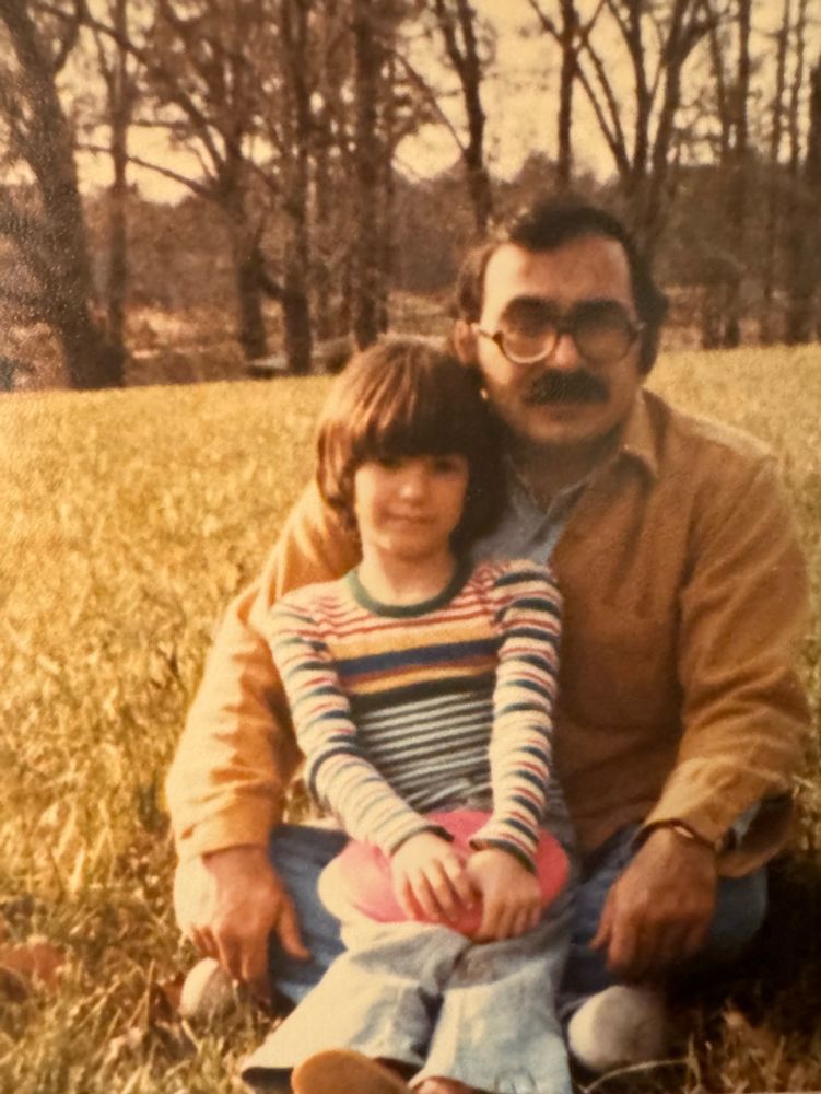 Old photo of a young girl with a haircut that looks like a bowl, sitting in a grownup's lap in a field, holding a frisbee
