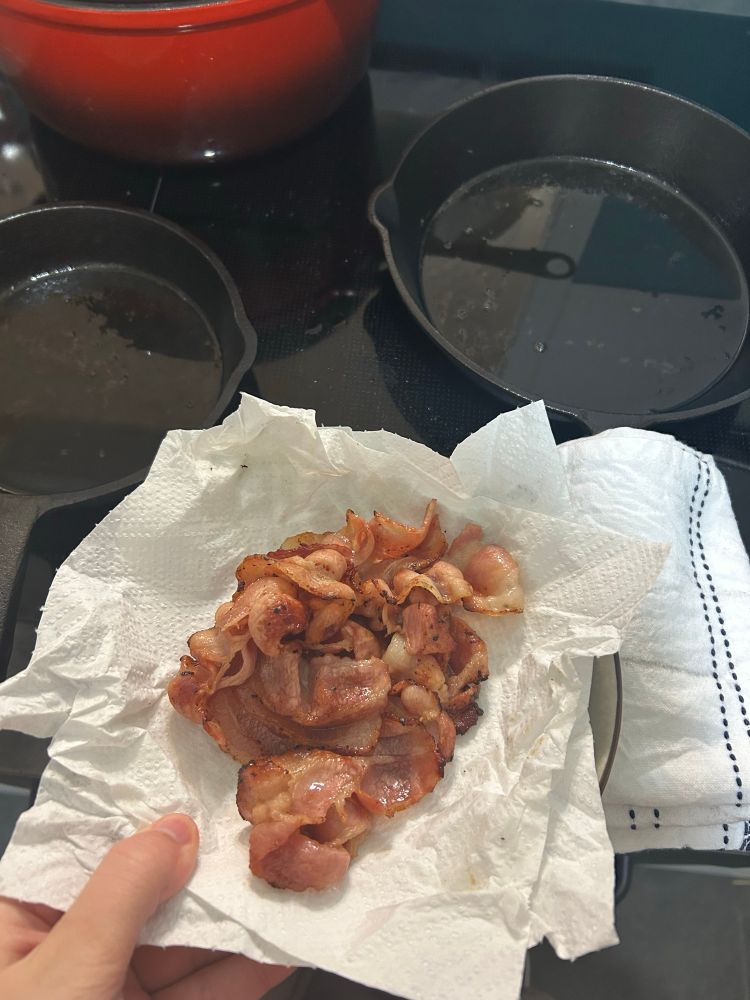 A plate full of bacon. Further back is a stove with two cast iron pans, shiny with pools of oil built up.