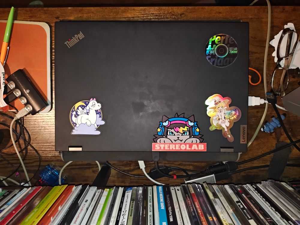 Lenovo Thinkpad on a desk with 5 stickers on it. Below the brand logo on the left is a cute dragon sticker from MOTEL777. Slightly right from center at the bottom is a Stereolab sticker, with the Ravefurrest sticker placed above it. At the top right is a holographic "Perfect Music Friday" sticker from PC Music. Below it is a holographic sticker from a Takashi Murakami gallery.