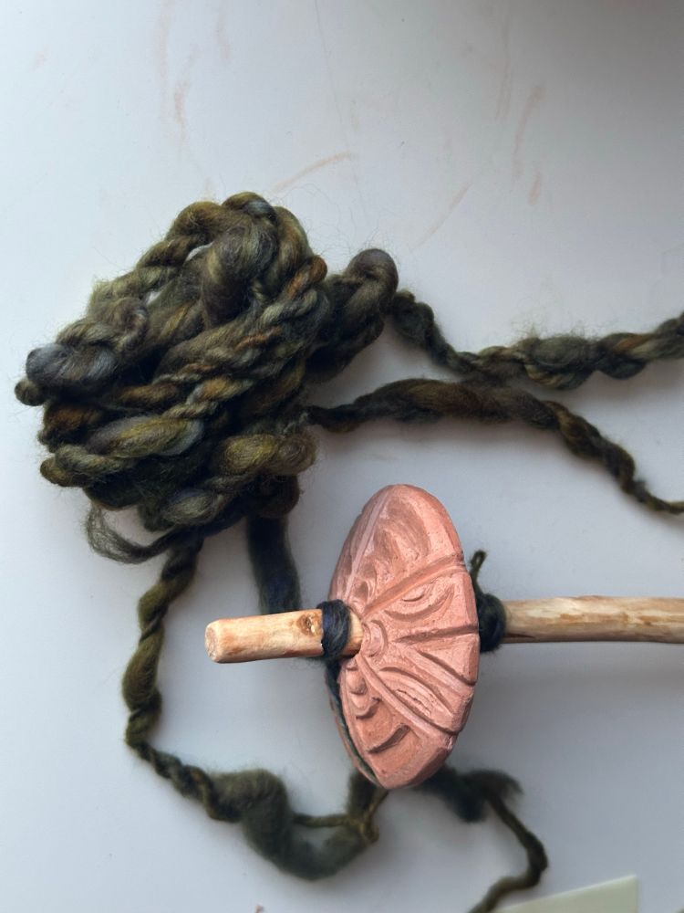 A photo of a rustic spindle with a clay whorl with a bundle of greenish-colored thick and thin yarn.