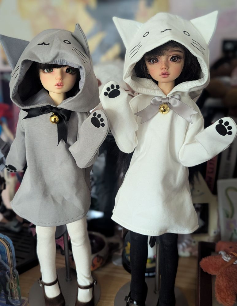 Two dolls dressed in dresses with cat hoods. The one on the left is in grey while the one on the right is in white.