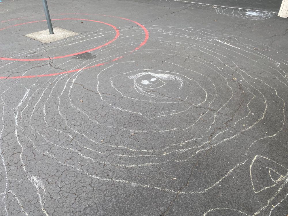 the asphalt floor of a playground with a chalk drawing of a crude face with an ambivalent expression surrounded by a 10 foot spiral