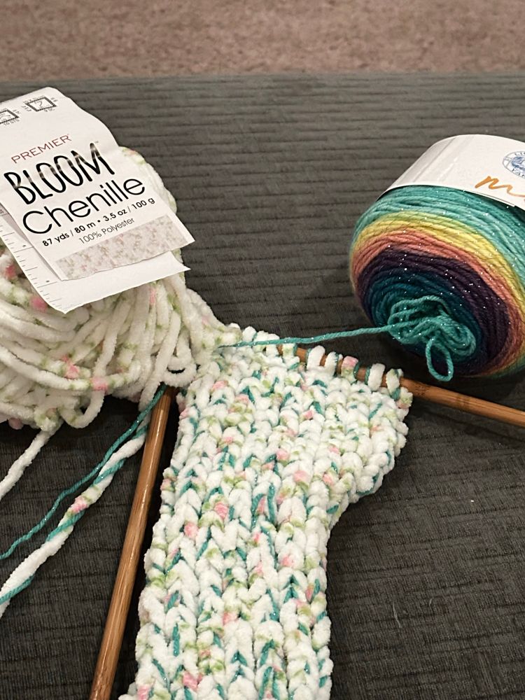 Two-stranded scarf on needles. Yarn is rainbow and a chenille with a subtle blossom pattern