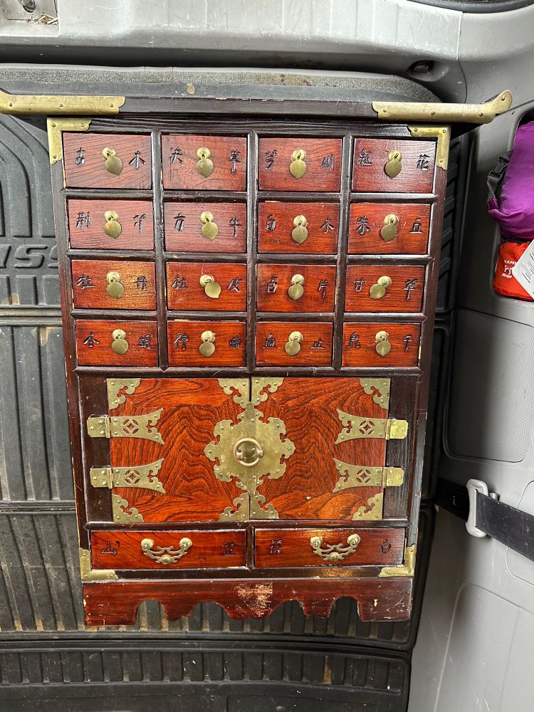 A narrow freestanding cabinet made of dark wood. Twelve mini drawers, one large open section, and two drawers at the bottom. It is fitted with brass-like catches and carved with Traditional Chinese characters for various herbs. 