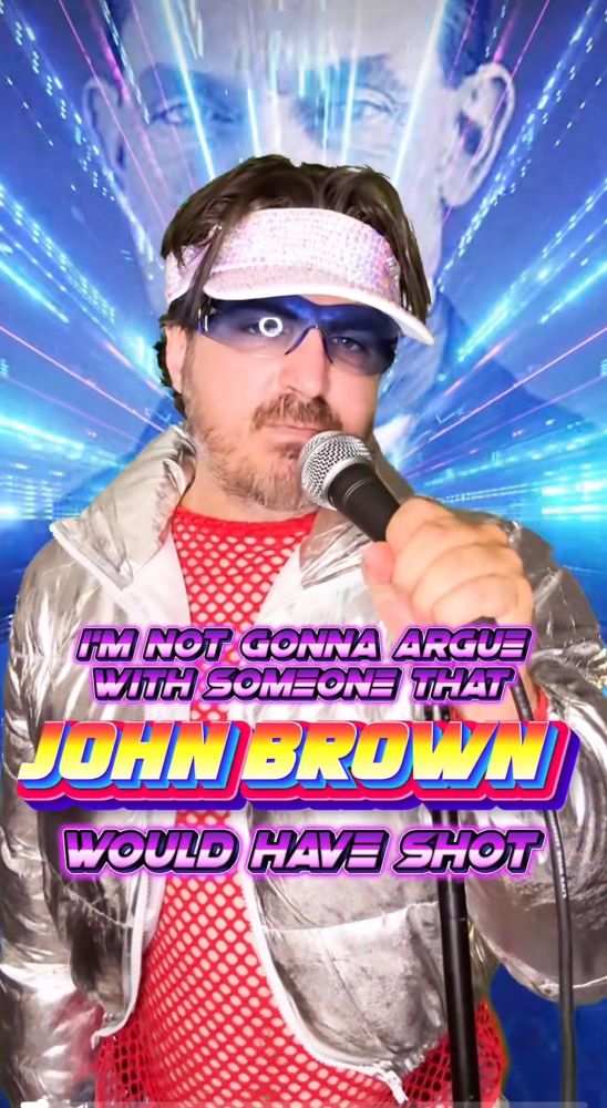 A man in a shiny metallic jacket holding a microphone in front of a laser background. The head of John Brown is superimposed behind him. The words “AM NOT GONNA ARGUE WITH SOMEONE THAT JOHN BROWN WOULD HAVE SHOT” are on the screen. 