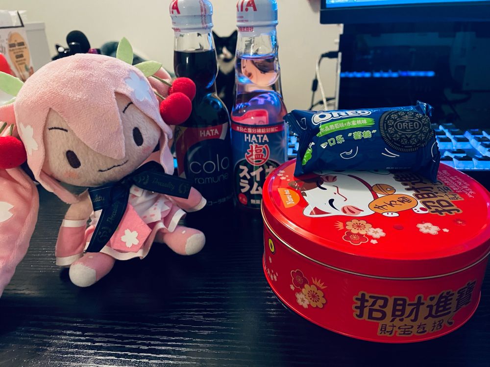 fuwafuwa sakura miku sitting on my black desk. cola and watermelon ramune next to her. tin of cookies and a packet of oreos at the other side.