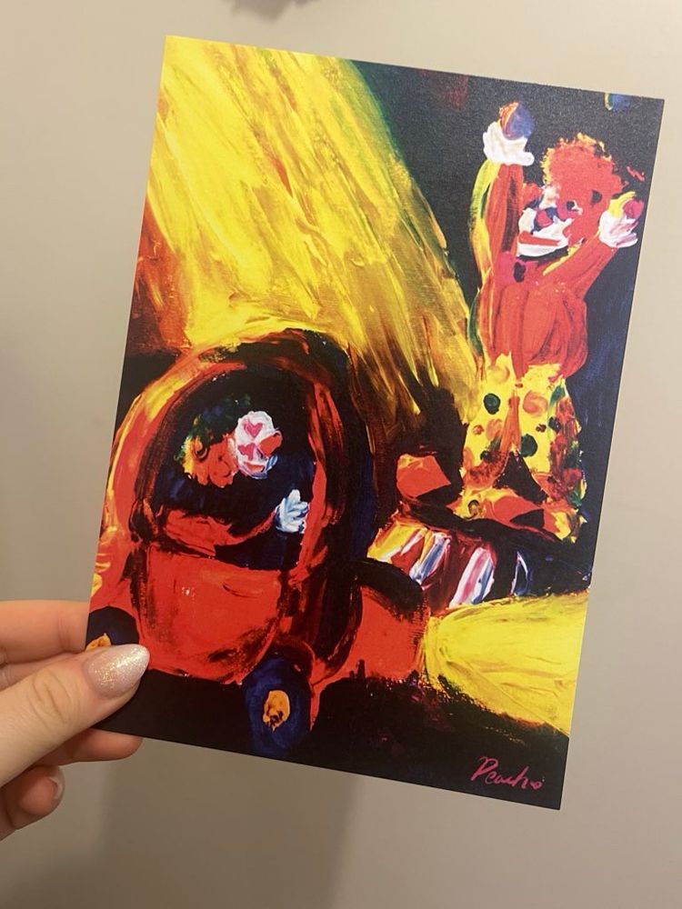 A hand holding a print of an acrylic painting, featuring a clown in a clown car looking with heart eyes at a clown on a stage juggling.