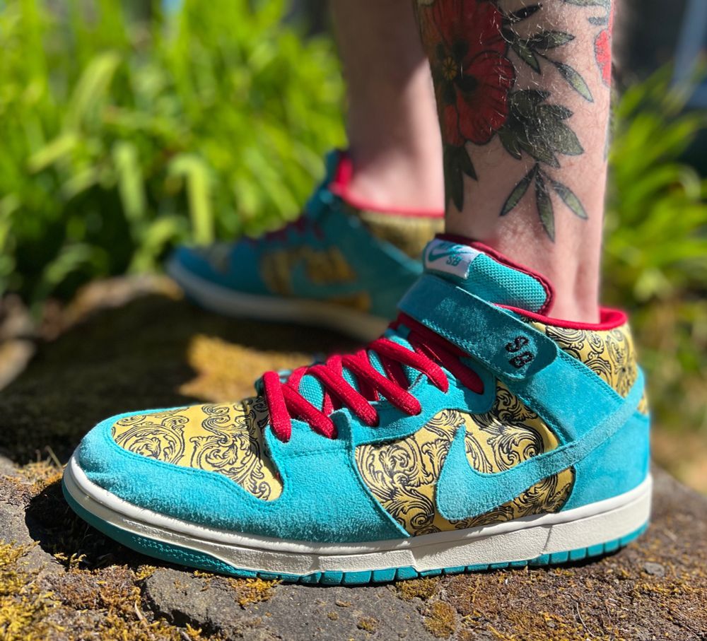 Nike Dunk Mid “Peacock” on foot outside. Light blue suede and gold floral brocade upper, red laces. Leg has floral tattoos.