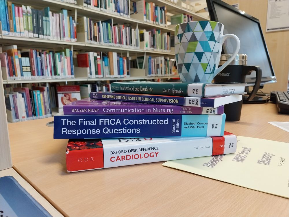A stack of medical textbooks sits on a desk with a brightly coloured mug balanced on top. The background shows a computer and shelves of books in a library.