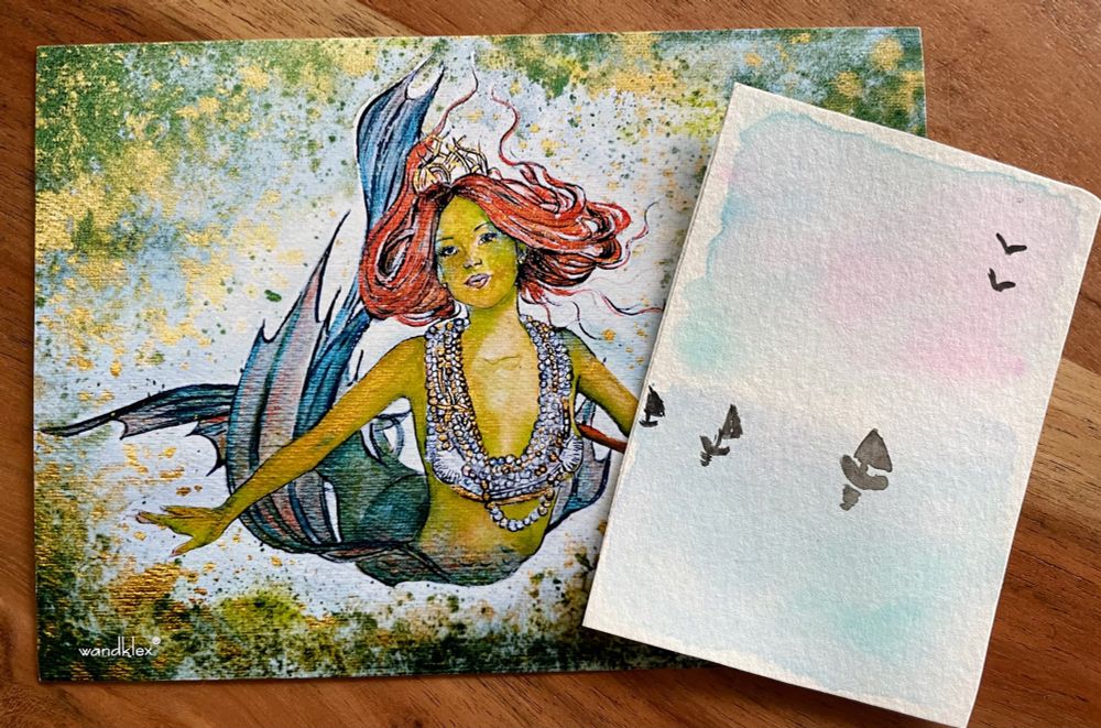 Eine ATC mit blau rosa Pastelltönen und schwarzen Segelbooten und Vögeln im Himmel. Dahinter eine Postkarte mit einer rothaarigen Meerjungfrau.