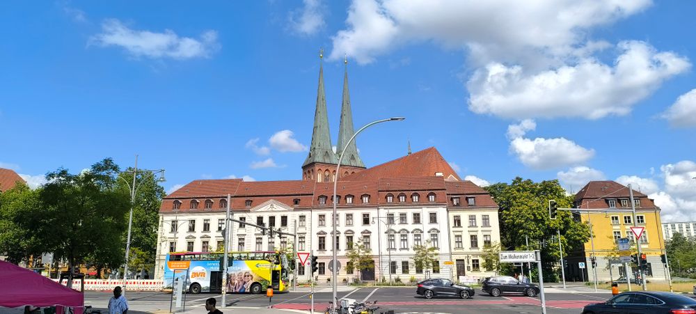 A big white building, two floors high, dominates centre stage, it has a red roof and just behind it are two green spires (I think that's what they are called, tall pointy things anyway) perhaps part of a church. It's a blue sky with lots of clouds and dark undersides. Location is Berlin, Alexanderplatz area, just opposite Alte Munze.