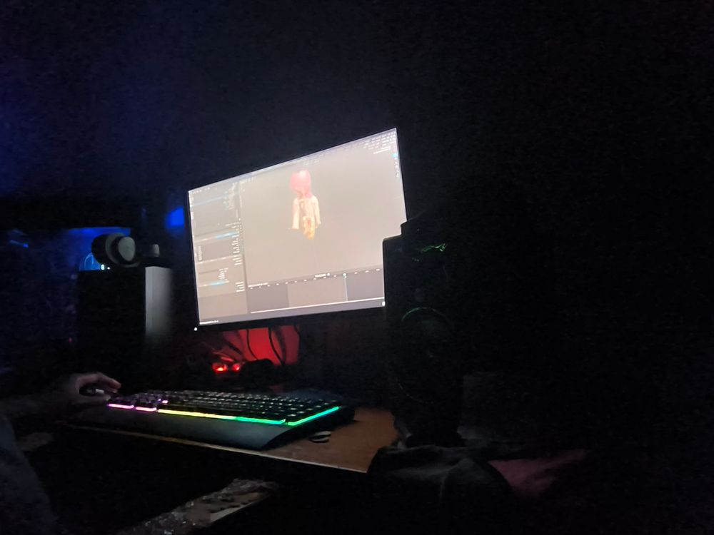Its hard to describe this photo, but it is of a desk with a computer screen on it, some 3D work on the screen, an RGB keyboad glowing, the desktop has blue lights in the background, I'm somewhere in the pic, barely visible.