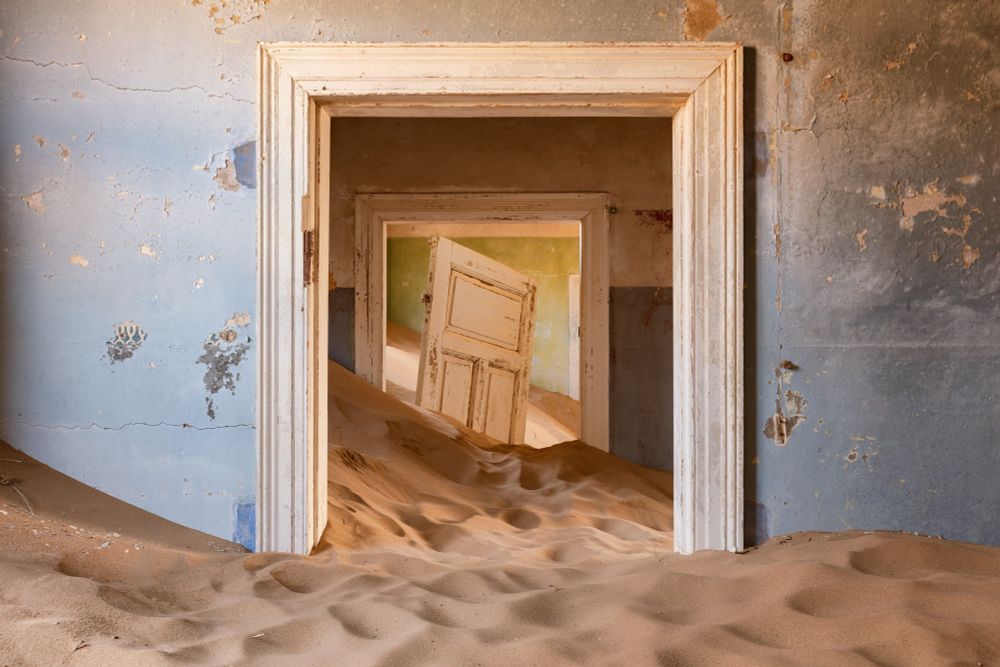 The view is through 2 doorways the rooms are full of drifted sand, this is an abandoned building - there is a door off its hinges in the centre of the second doorway.  The walls are a powder blue almost looking like the colour of the sky against the sandy floors
