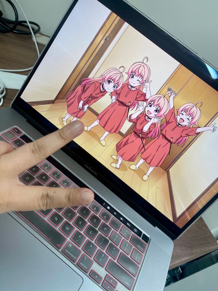 A hand flashes a peace sign in front of a laptop screen showing a scene from the anime Witch Watch, where four duplicated versions of the character Nico stand together in red outfits inside a hallway.