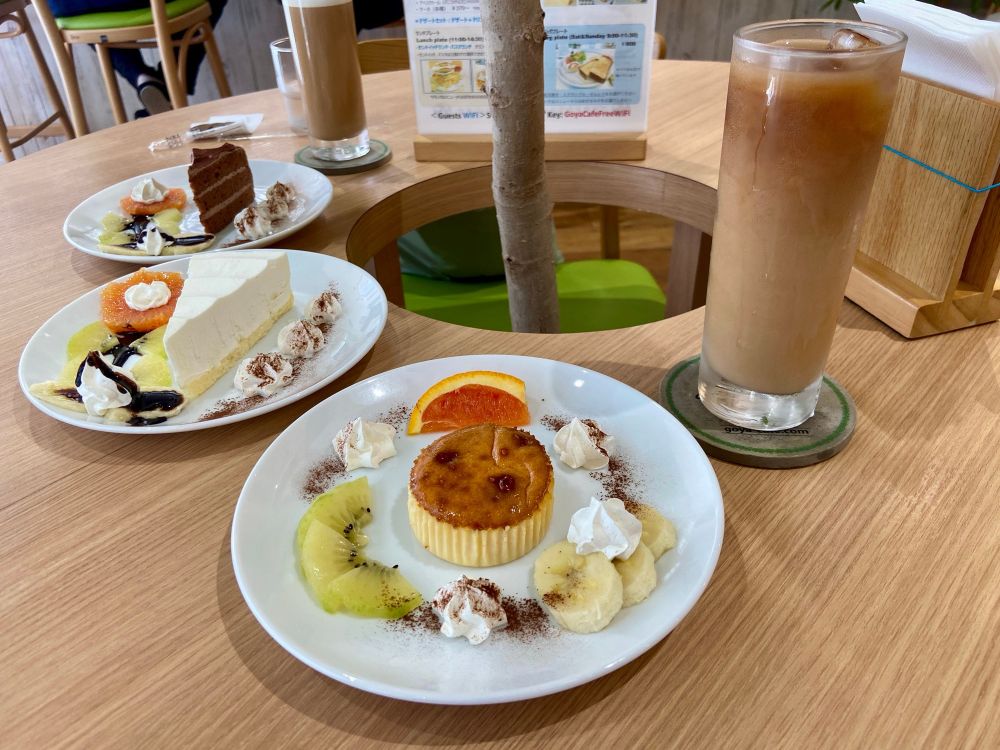 Café table with plates of desserts including cheesecake, chocolate cake, fruit, and a brûlée tart, along with an iced latte.