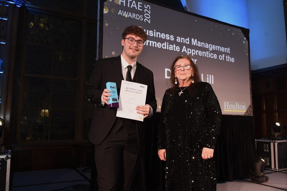 HTAE Apprentice of the Year, Declan Lill, with Cllr Linda Tock. They both wear smart black clothes and pose in front of a large screen that announces Declan's win.