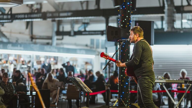 A man holding a guitar sings into a microphone inside Trinity Market in Hull, in front of a crowd of people sat in chairs and on benches. 