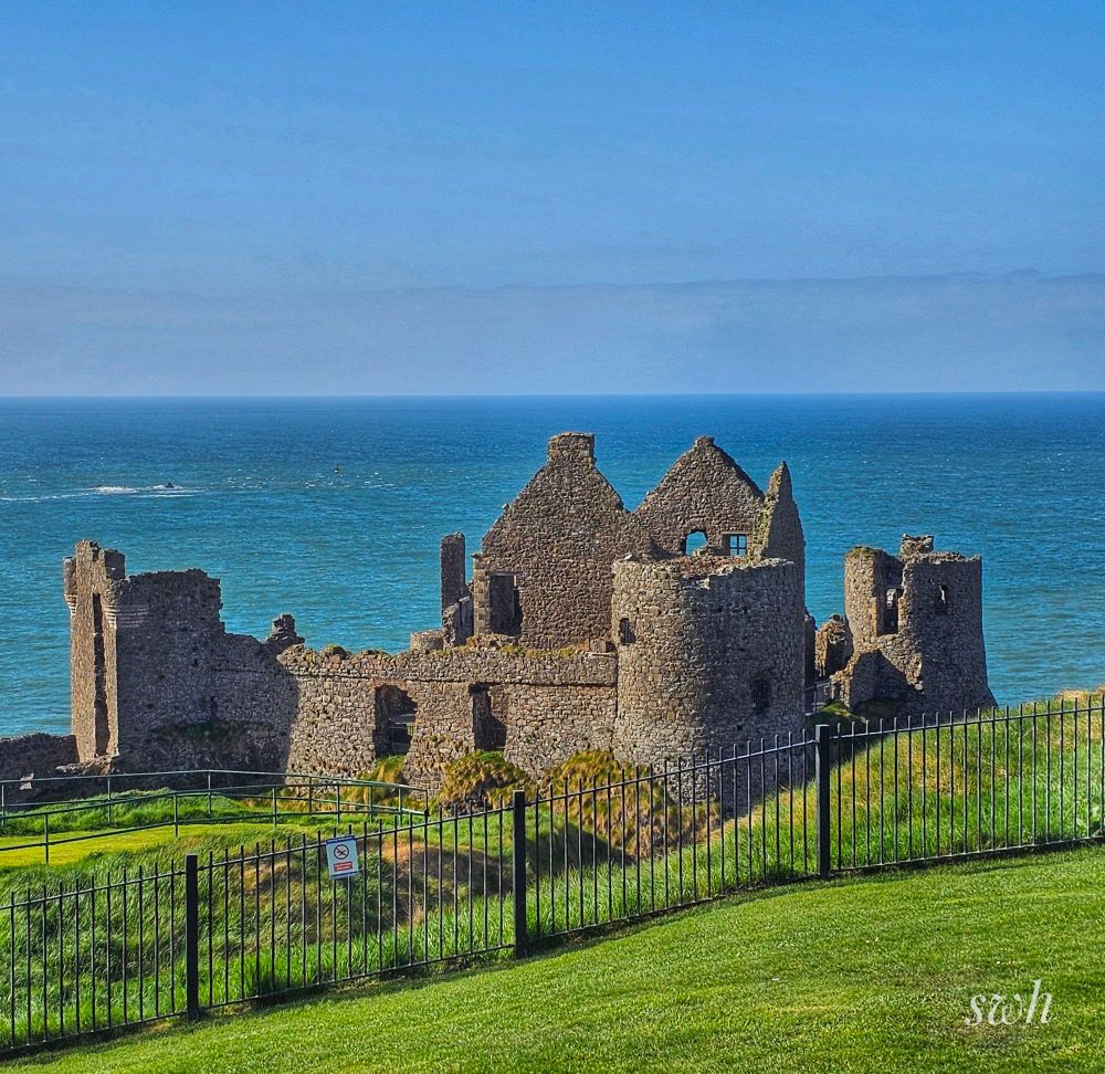 Grass with a metal fence in the foreground, with the ruins of a castle perched close to the edge of a cliff with the sea beyond it.