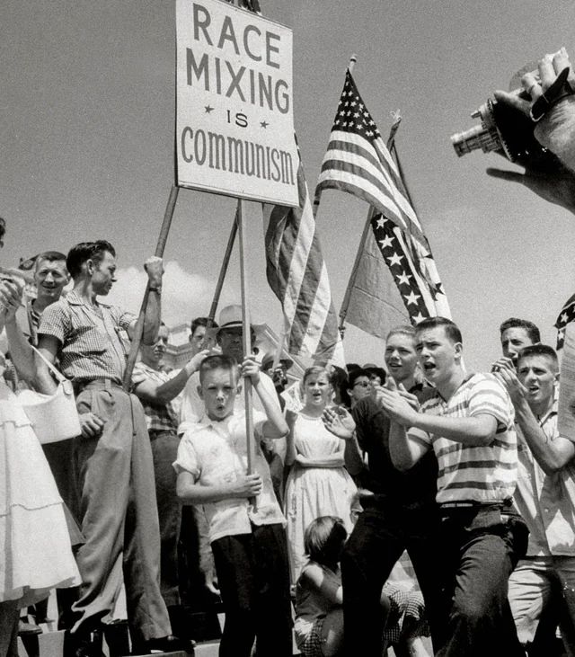 White racists in America carrying signs and waving Confederate flags to protest integration in 1954. A young boy holds a sign that reads “Race mixing is Communism.” Women, boys, and men—but mostly boys and men—clap their hands and appear to be singing. Just out of frame, a reporter points a movie camera at the assembled protesters. 