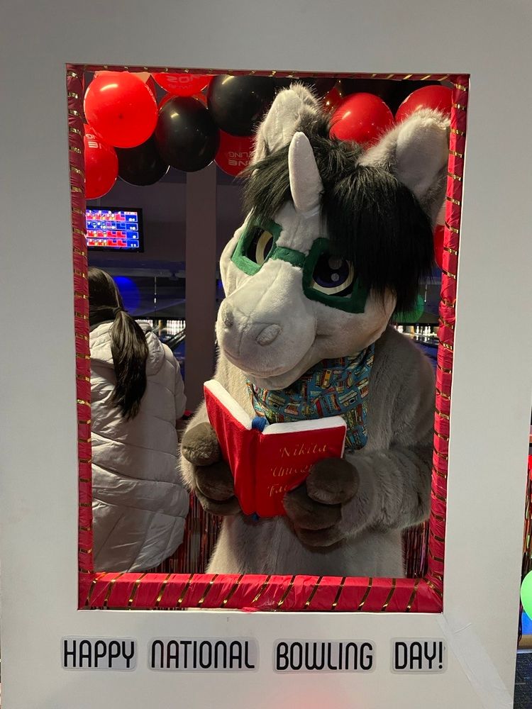 A unicorn fursuiter holding a book, looking through a frame that says Happy National Bowling Day