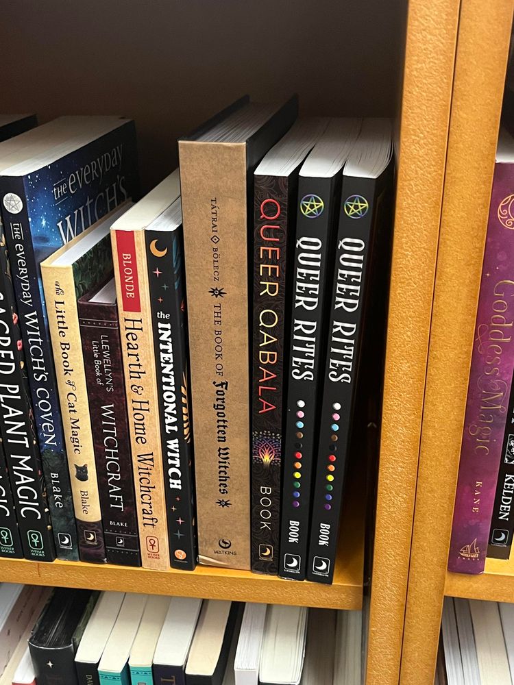 A set of books on a shelf at a bookstore. On the far right of the shelf are Queer Qabala and two copies of Queer Rites, both by Enfys J. Book.