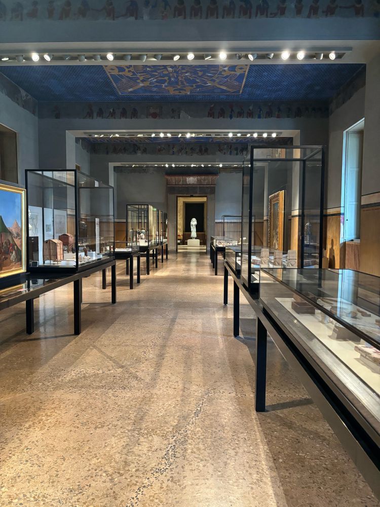 Picture in one of the long room in the Egyptian museum and papyrus collection in Berlin. You see some showcases with objects on the left and right side. The end of the corridor leads to a statue of a pharaoh. The ceiling is blue and shows a zodiac