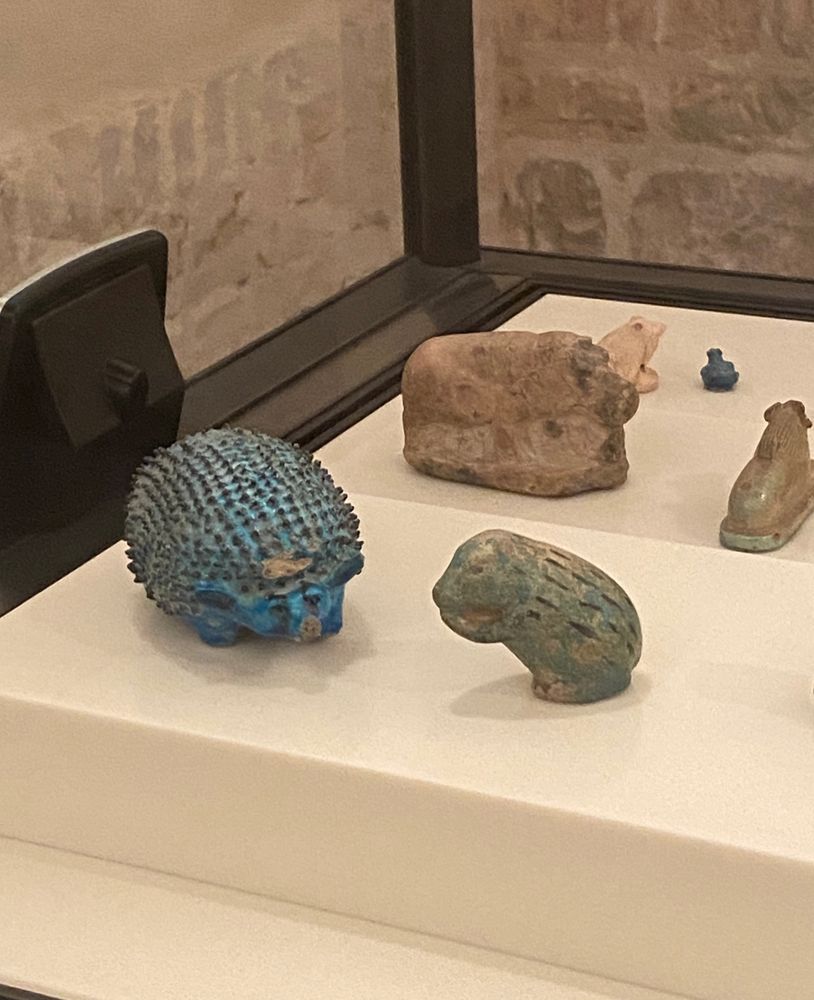 Picture of two animal figurines from ancient Egypt on a museum display. The figurines depict a hedgehog and a shrew, both made of faience. In the background: a few more animal figurines