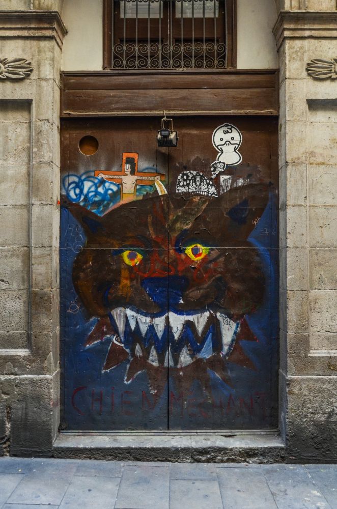 Un graff sur une porte d'un squat à Barcelone. l'image représente un chien aggressif en grand, avec quelques dessins abstrait ou picturaux au dessus en petit. Il y a une inscription "Chien méchant" en dessous. (photo prise en 2015)
