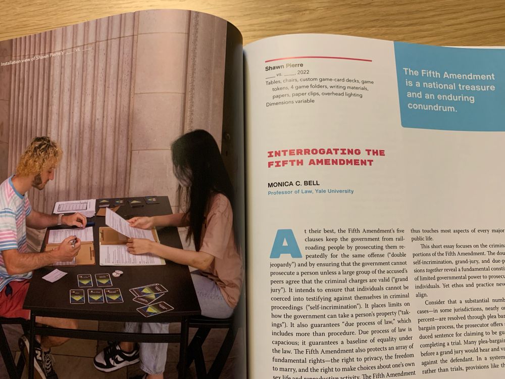 A photo of the inside of a book. The left side features two people playing a tabletop game, the right text about the exhibition piece.