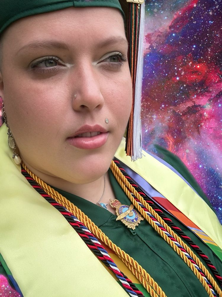 Portrait of a graduate wearing a green cap and gown. They have white and multi-colored tassels and honors cords adoring their neck. There is a yellow highest honors stole around their neck, too. They have a blue Topaz Star Trek necklace and a Royal Air Force clip on their zipper. The background is a faux purple and pink nebula. The person is looking off into the distance pensively. They have green eyes and pink glossy lips, and they have two gold facial piercings. And they are wearing scarab pearl earrings.