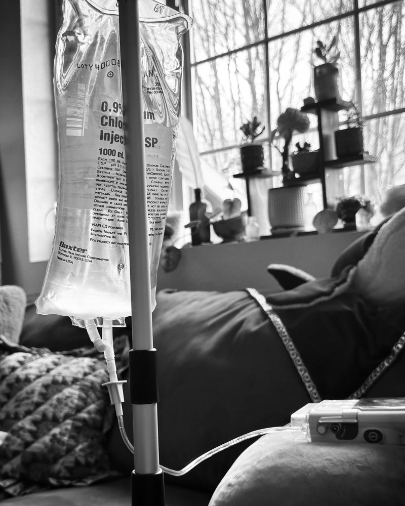 Black and white photo of an IV pole and IV bags for chemotherapy. The background is a home with a couch and pillows. There is a window above the sofa with many small potted plants on it.