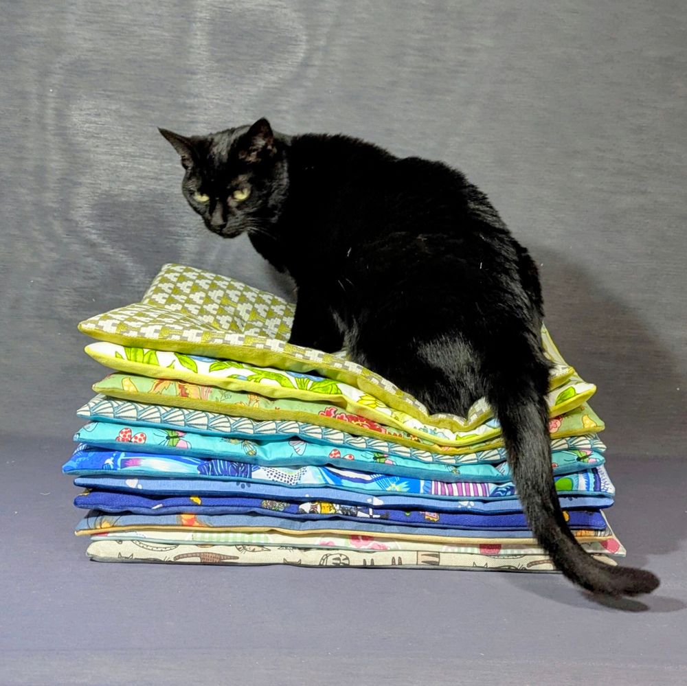 A black cat is sitting on top of a pile of lap mats in various colours .. mostly blues and lime greens 