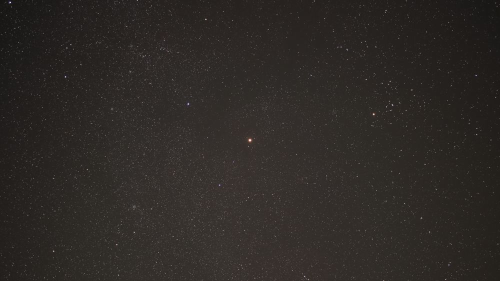Bright orange planet Mars in the centre of a field of stars on the night of 2 October 2022. Mars is within the constellation of Taurus, between the stars of the 'horns of the bull', Elnath and Tianguan. The Hyades open cluster can be seen to the right of Mars, with the bright orange star Aldebaran prominent. Equipment and settings: Canon 600D with 24mm lens on Star Adventurer mount. Stacked shot of 13x 30s exposures at 24mm fl, f/2.8, ISO 1600.