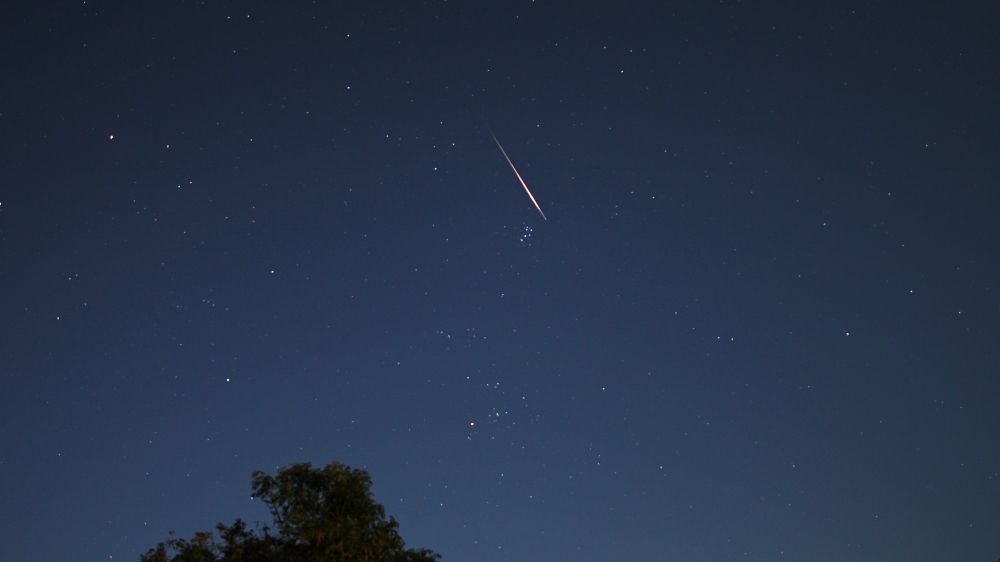 A green and orange Perseid meteor captured in the constellation of Taurus. It appears above the Pleiades open cluster. The sky is a deep blue colour due to moonlight. A tree is in the foreground. Settings: 8s exp, 16mm fl, f/2.0, ISO-400.