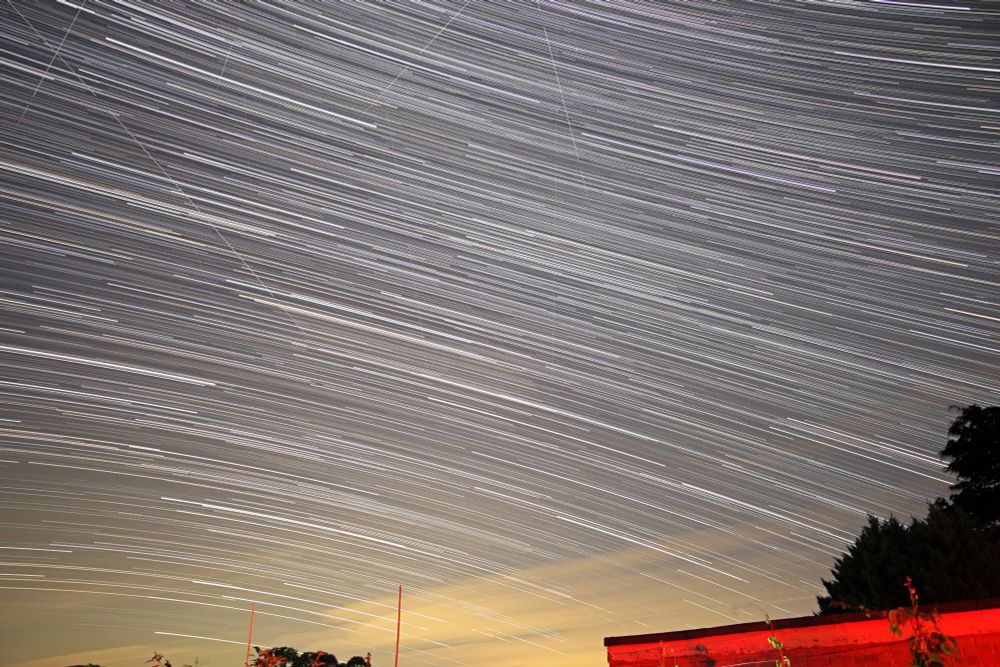Streaks of light arcing across the night sky with a foreground of trees and buildings. A star trails shot covering 1 hour 15 minutes of star movement, centred on the constellation of Scutum.