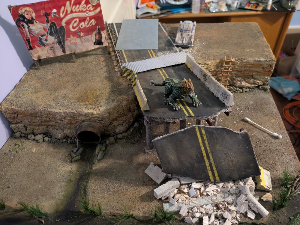 A cliff side scene with a collapsed overpass, a weathered Nuka Cola bill board. A nasty drain empties into a foreground bog. Colored in Southwest pallette with various shades of iron in rock. An abandoned Corvega sits in the background.