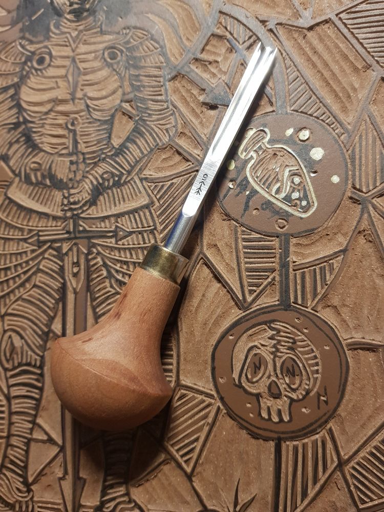 Photo proche d'une gouge posée sur une plaque de linogravure marron. On peut deviner sur les motifs une femme en armure, un crâne et une potion