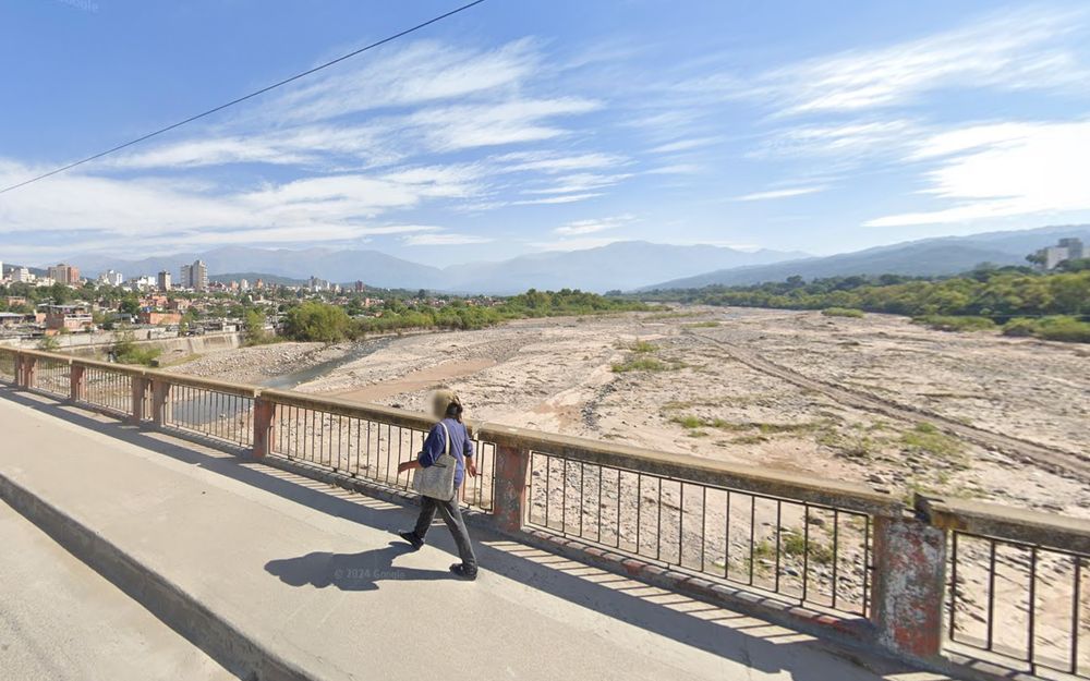 Google Street View screenshot: a person walks along a bridge over a wide braided riverbed, San Salvador de Jujuy, Argentina. In the background, hazy mountains loom over a cityscape dotted with small towers.