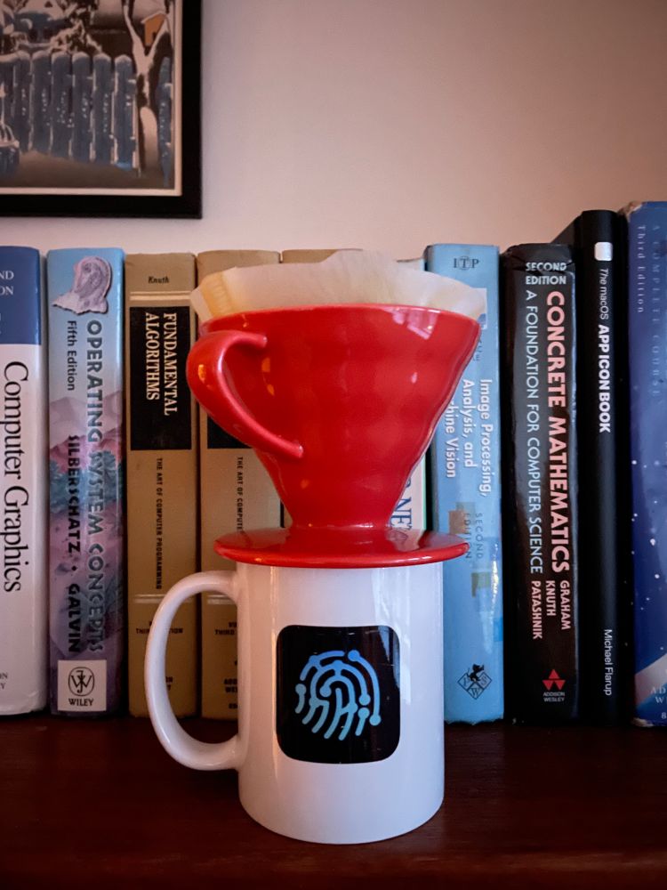 Coffee mug with Working Copy icon and dripper on top standing in front of technical books by Donald Knuth and others.