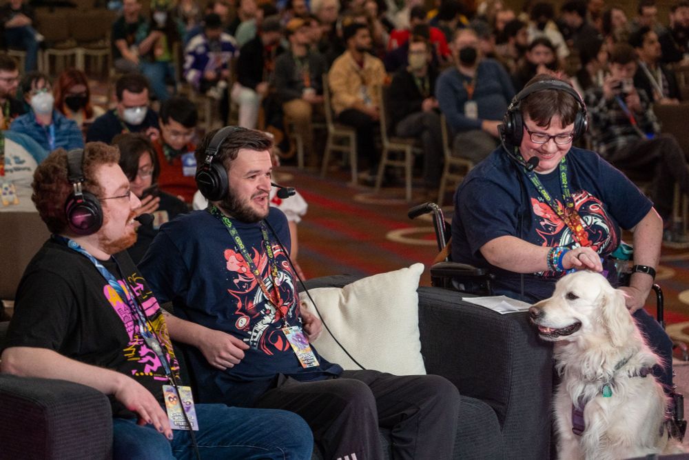 Two people sit on a couch while a third person sits in a wheelchair and a white golden retriever sits in front of the person in the wheelchair. All three people are wearing large headsets and there is a crowd sitting in chairs behind them that is blurry.
