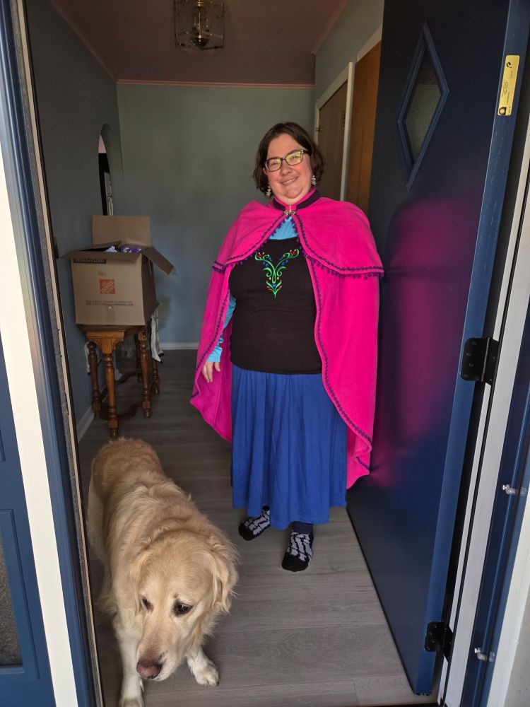 A photo through a doorway. An adult femme stands in an outfit inspired by Anna from Frozen with a pink cape, light blue undershirt, black outer shirt with a green vine pattern, a dark blue skirt, and skeleton socks. A white golden retriever stands to the person's right, looking out the door. 