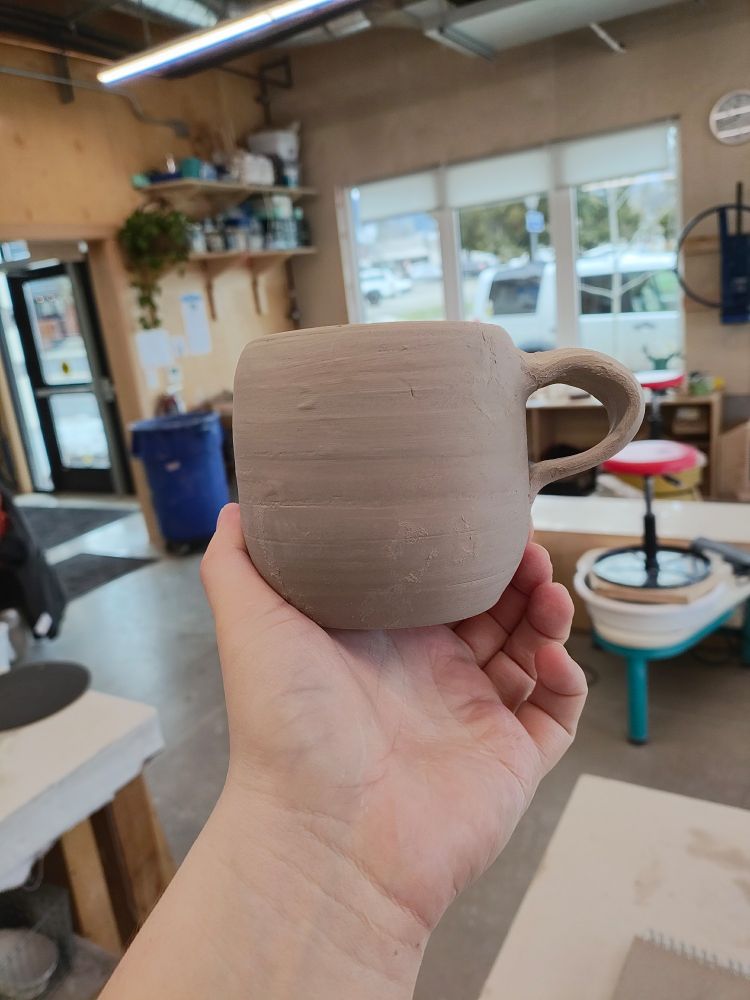 A trimmed mug with a pleasantly curved bottom is held aloft in a pottery studio. A misshapen handle sticks out from the side, starting just below the rim and curving out and down to attach about halfway down the mug