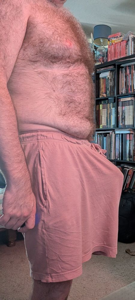 A hairy man wearing pink boxer shorts stands in frame, something underneath his shorts stands at attention, causing a distinct tenting.