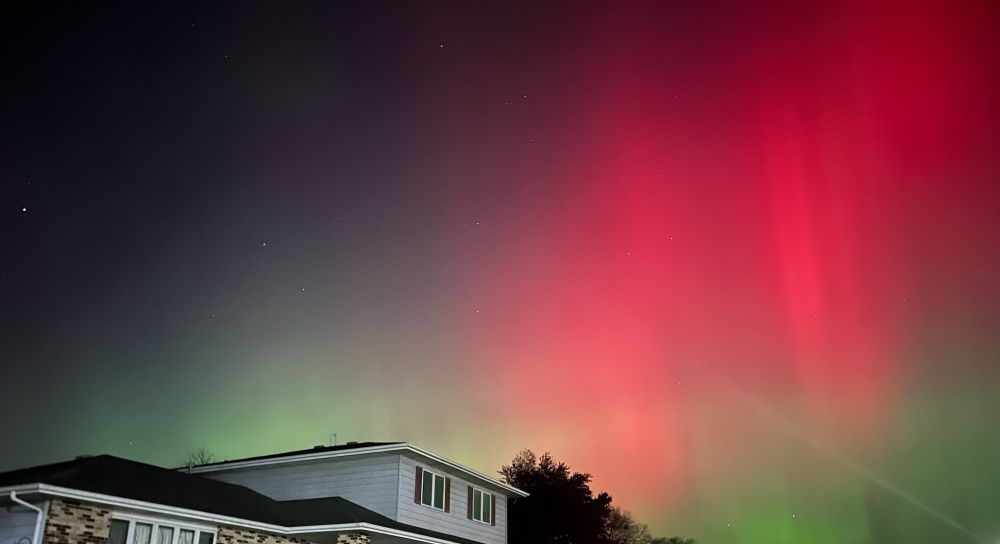 pic of the rooftop of a house - the northern lights fill most of the sky: beautiful green and vivid red with a black starry heaven beyond. 