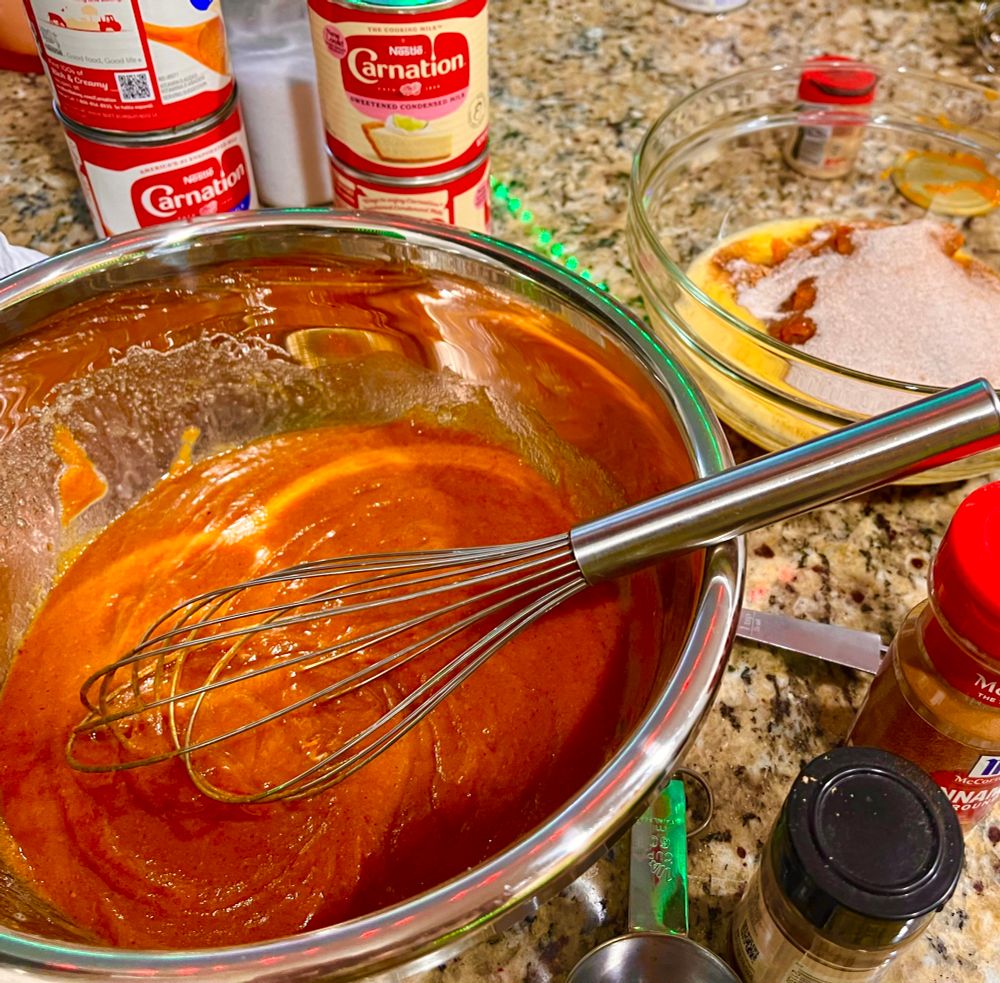 A bowl with half-finished pumpkin pie filling and many spices around the edges. 