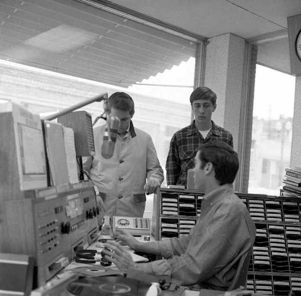 A dj seated at his console talks to two boys on the other side of the desk. One wears a coat, the other a plaid Pendleton shirt.