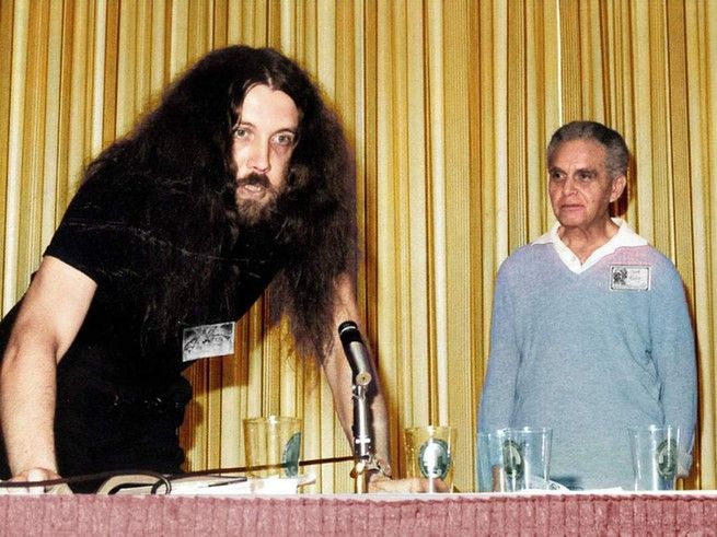 Alan Moore and Jack Kirby at a convention, standing behind a table. Jack stands back as Alan leans on the table over a microphone, mouth open, eyes unfocused