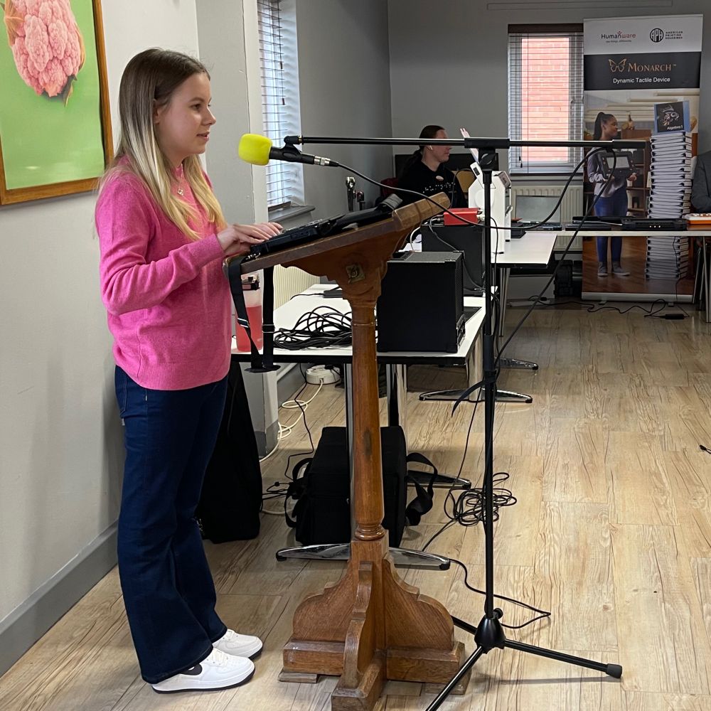Eleanor is standing at a wooden podium, speaking into a microphone with a yellow foam cover. She is wearing a bright pink jumper, dark blue jeans, and white trainers. Her long blonde hair is down. Behind her, there is a table with audio equipment and cables. The room has light-colored wooden flooring and gray walls, with a window partially visible. A framed picture of a pink flower is hanging on the wall to the left.