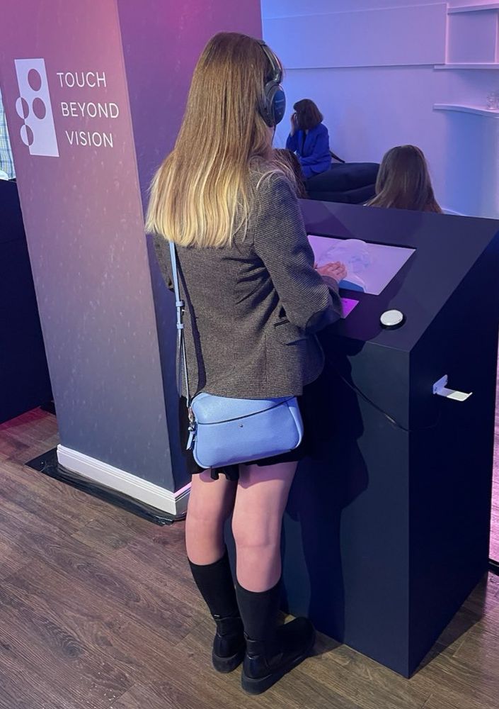 Eleanor is standing with her back to the camera, facing a dark black kiosk. She has long, straight, light brown hair with blonde highlights at the end. She's wearing a brown blazer, a black dress, and black knee-high socks with black boots. A light blue purse hangs on her left side. She has large, black over-ear headphones on. Her hands are on a raised, white textured surface on the kiosk. To the left of her the supporting beam to the room is adorned with a representative braille graphic logo that also reads “TOUCH BEYOND VISION". The lighting in the room is a mix of purples and pinks.