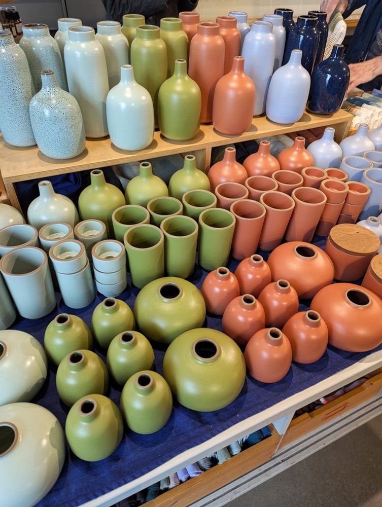 Photo of colorful vases from the Heath Ceramics shop. Rows of tomatillo green, salmon, and eggshell baby blue bud cases, round vases, and tall vases sit side by side on a wooden shelf.